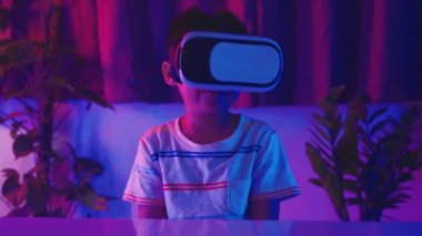 Sanal gerçeklik gözlüğü takan Asyalı küçük çocuk gerçeklik gözlüğü takıyor, çocuk VR başlığı takıyor sola bak, sağa bak ve evde heyecanlı ol koyu mor ve mavi arka plan, sanal teknoloji.
