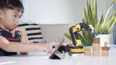 Asyalı küçük çocuk atölyede dijital tablet bilgisayar kullanıyor, çocuk ders programlama kontrol robotu kolu öğreniyor tahta bloğu kaldırıyor, teknoloji bilimi eğitimi alıyor.