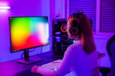 Masaüstü bilgisayarında çevrimiçi video oyunu oynayan Asyalı profesyonel oyuncunun renkli neon ışıklı karanlık odası, oyun kulaklıkları takan genç kadın evde oyun oynamak için bilgisayar kullanıyor, Arka Görüş