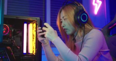 Oyun kulaklığı takan Asyalı kadın oyuncu video oyunu oynamak için joystick konsolu tutuyor ve evdeki bilgisayar PC neon ışığı ile çevrimiçi yayın yapıyor, turnuva oyuncusu E-Sport konsepti