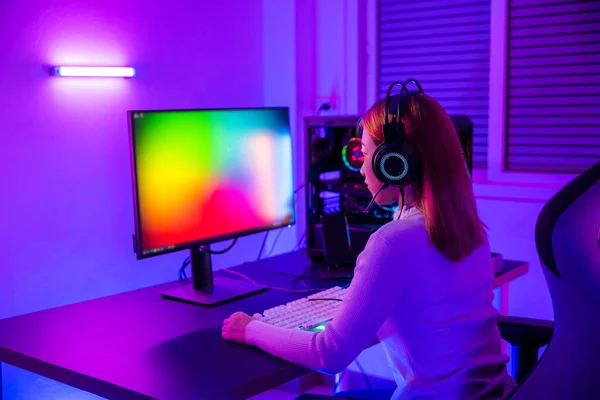 Asian professional gamer playing online video game on desktop computer PC have colorful neon LED lights, young woman in gaming headphones using computer for playing game at home