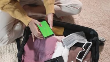 Woman using smartphone blank green screen and packing suitcase bag for travel trip weekend vacation at home, female check information in tourism website on mobile phone before go to airport