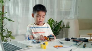 Asyalı çocuk tekeri Arduino robot araba ev ödevine monte ediyor, küçük çocuk tekerleği araba oyuncağına monte etmeye çalışıyor, elektronik yapay zeka atölyesi online okul dersi yaratıyor.