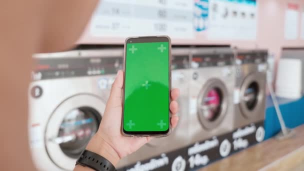洗濯機作業中に洗濯機でデジタルスマートフォンを使用する女性 セルフサービスの洗濯設備 スマートフォンの緑の画面で洗濯するための女性の設定アプリケーション — ストック動画