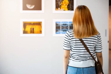 Asyalı bir kadın renkli çerçeveli resimlerin önünde dikiliyor beyaz duvarda resimler, genç bir kadın fotoğraf çerçevesinde sergi galerisine yaslanıyor,