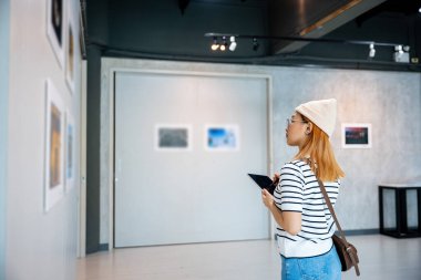 Asyalı kadın sanat galerisindeki tablete bilgi not alıyor. Çerçeveli resimlerin önünde duvarlara bakıyor. Fotoğraf çerçevesindeki genç kişi sergi sanat galerisinde dijital kitap yazıyor.