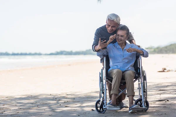 Happy Asian elderly woman sitting in wheelchair and husband is a wheelchair user smartphone taking selfie on the beach, summer vacation, Retirement couple concept