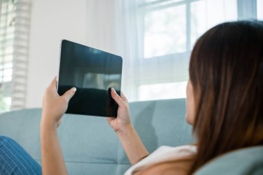 Mutlu Asyalı kadın evinde akıllı tablet oynuyor kanepe oturma odasında dinlenirken, güzel kadın kanepeye oturmuş dijital tablet kullanıyor, sosyal medya teknolojisi kullanıyor.