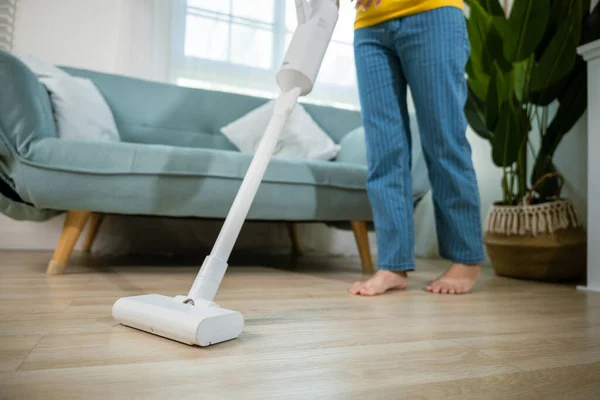 Happy Asian young woman with accumulator vacuum cleaner at home in living room, housewife female dust cleaning floor under sofa or furniture with vacuum cleaner, household and housework concept