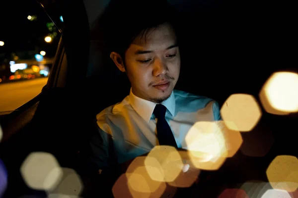 Businessman using smartphone in backseat of car near window at night, Handsome Asian young man work on late night sitting back seat and still work with mobile phone in city street