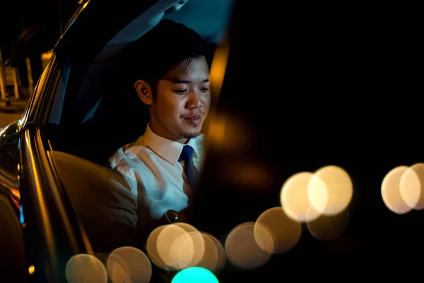 Businessman using smartphone in backseat of car near window at night, Handsome Asian young man work on late night sitting back seat and still work with mobile phone in city street