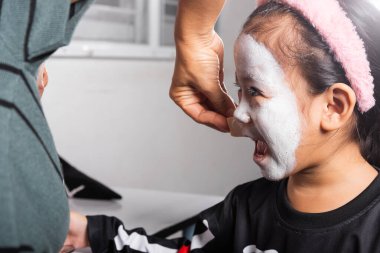 Asian mother and kid daughter getting makeup halloween face painting look like ghost, Portrait of woman applying color brush to child girl face for party, Happy halloween day concept