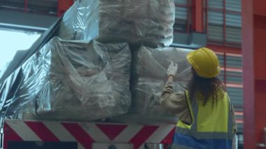 Endüstriyel kadın kıvırcık saç mühendisi, depo atölyesi, lojistik ve taşımacılık atölyesindeki müşteriye sevkiyattan önce kargo kamyonunda metal levhaları kontrol etmek için güvenlik üniforması giyiyor.