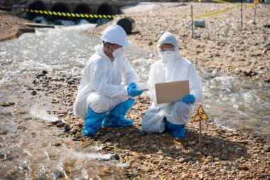 Ekolojist ölü balığı incelemek ve dizüstü bilgisayara veri kaydetmek için örnek alıyor, biyolog koruyucu giysi giyiyor ve endüstriden, sorun ortamından gelen atık su örneklerini maskeliyor