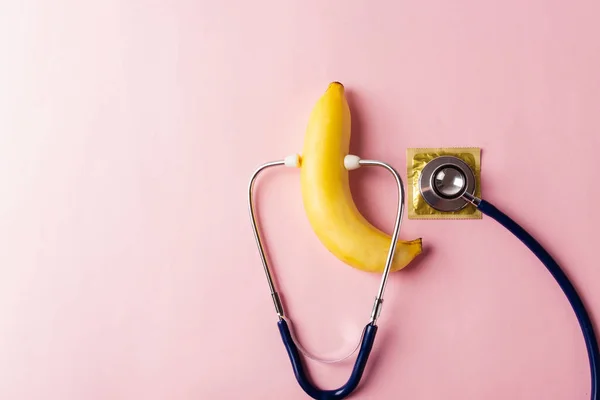 World sexual health or Aids day, Top view flat lay condom in wrapper pack, banana and doctor stethoscope, studio shot isolated on a pink background, Safe sex and reproductive health concept