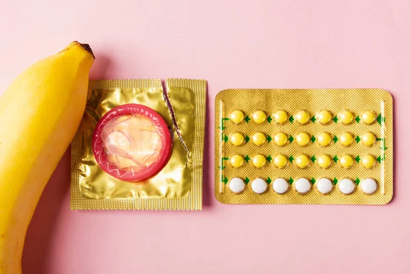 World sexual health or Aids day, Top view flat lay condom on wrapper pack, banana and contraceptive pill, studio shot isolated on a pink background, Safe sex and reproductive health concept