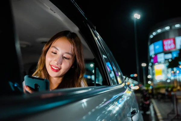 Asian businesswoman commuting from office in Taxi backseat with mobile phone on road in city at night after late work, Beautiful woman using smartphone sitting back seat her car in urban