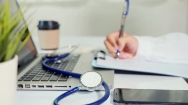 Closeup of doctor or nurse woman in uniform with stethoscope writing information of patient prescription in paperwork on clipboard and typing laptop computer for history record medical document report