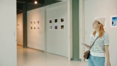 Asian woman walking through art gallery collection in front framed paintings pictures on white wall, Visitor person walk looking to picture at photo frame at interior show exhibition artwork gallery