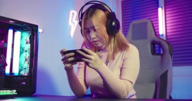 Gamer playing has aches and pains twisting left and right while playing in online video game long times with on smart phone with neon lights, Asian woman wearing gaming headphones play smartphone