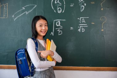 Back to school. Portrait of smiling woman child student of black chalk board, Happy beautiful Asian Schoolgirl girl standing holding books standing in front blackboard of classroom, Education lesson