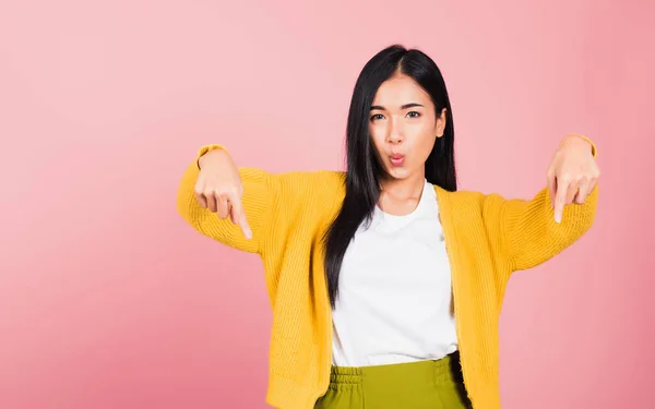 Asian happy portrait beautiful cute young woman smiling stand makes gesture two fingers pointing below down presenting and looking to camera, studio shot isolated on pink background with copy space