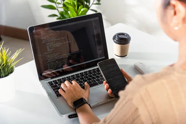 Programmer woman looking mobile phone program code at workplace, software developer hands typing data coding programming javascript with laptop computer, IT startup concept, Web application design