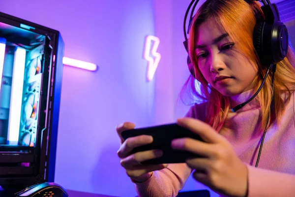 Happy Gamer playing video game online with smartphone with neon lights, Asian woman wearing gaming headphones play game on mobile phone at home, E-Sport concept