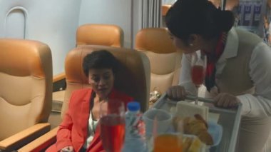 Charming stewardess in uniform flight attendant walking on aisle serve food and service to woman passengers on his cart in airplane, air hostess cabin crew service inside aircraft passenger seat