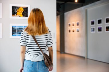 Asian young woman standing she looking art gallery in front of colorful framed paintings pictures on white wall, female watch at photo frame to leaning against at exhibit museum, Back view
