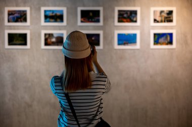Photographer visit at photo frame to leaning against at show exhibit artwork gallery, Asian woman takes picture art gallery collection in front framed paintings pictures on white wall with camera