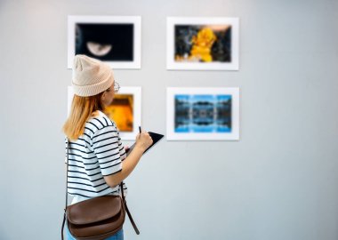 Young person at photo frame write digital book leaning against at show exhibit artwork gallery, Asian woman holding tablet at art gallery collection in front framed paintings looking pictures on wall
