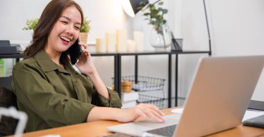 Happy Asian young woman working on laptop computer at home office while talking on mobile phone, smiling businesswoman having business call to talking with company sales client financial