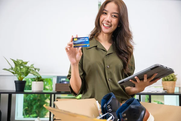 Asian young woman received online shopping parcel opening shoes boxes to checking on tablet, Cheerful woman holding credit card, happy female unpacking cardboard box, writing online store feedback