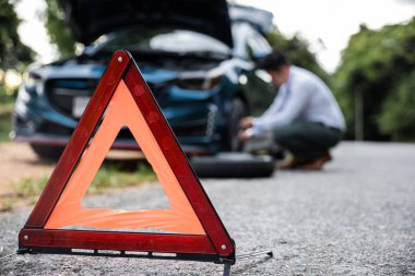 Asian businessman car broken has problems with wheel of his car during go to work in morning he changing replace rubber tire on wheels, business man have warning sign on road, problem transportation