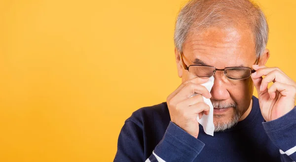 Portrait Asian senior old man sad wiping away his tears studio shot isolated on yellow background, Elder man crying raise glasses with tissue wipe red eyes, Sadness depressed lonely