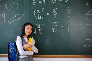Back to school. Happy beautiful Asian Schoolgirl girl standing holding books standing in front blackboard of classroom, Portrait of smiling woman child student of black chalk board, Education lesson