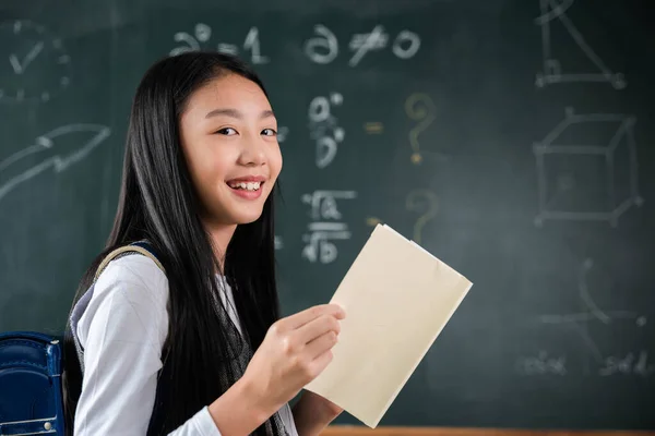 Back to school. Happy beautiful Asian Schoolgirl girl standing holding books standing in front blackboard of classroom, Portrait of smiling woman child student of black chalk board, Education lesson