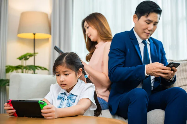 Family don\'t care about each other. Asian parents ignore their child and looking at their mobile phone at home, father and mother read social media but daughter play video game on sofa living room