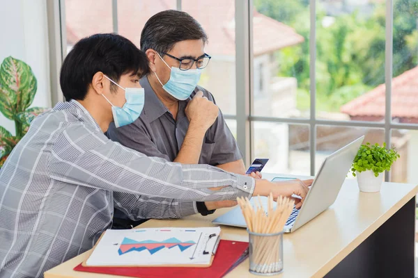 Asian young man teaching senior father how to use credit card for shopping online with laptop computer in the living room, adult son holding bank card helping Dad to purchases web online application