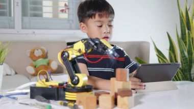 Asyalı küçük çocuk atölyede dijital tablet bilgisayar kullanıyor, çocuk ders programlama kontrol robotu kolu öğreniyor tahta bloğu kaldırıyor, teknoloji bilimi eğitimi alıyor.