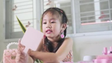 Asyalı sevimli küçük kız makyaj yapıyor ve aynada kendine bakıyor, kadın olmayı öğreniyor, mutlu çocuk güzel bir makyaj oyuncağı.