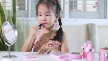 Asyalı sevimli küçük kız yüzünü makyaj yapıyor aynaya bakıyor ve dudaklarına kırmızı ruj sürüyor, kadın olmak için etkinlik öğreniyor, mutlu çocuk güzel makyaj yapıyor kozmetik oyuncağıyla.