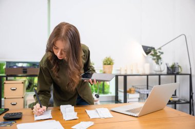 Finansal verilerle çalışan Asyalı genç bir kadın tablet bilgisayar ve yazma ile hesaplama yapıyor, hesaplama yapıyor, aile bütçesini ve muhasebeyi hesaplayan bir kadın, faturaları çevrimiçi ödüyor.