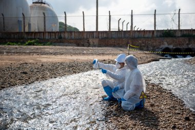 Nehirden çıkan zehirli kimyasalları test tüpü camlarıyla test eden ve beyaz duman çıkaran ekolojist, koruyucu kıyafet giyiyor ve maske endüstriyel, sorunlu ortamdan örnek su topluyor.