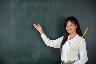 Back to school concept. Happy beautiful young woman standing hold pointer to back board, Asian female teacher smiling with wooden stick pointing to blackboard at school in classroom, Education