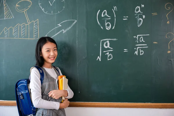 Back to school. Portrait of smiling woman child student of black chalk board, Happy beautiful Asian Schoolgirl girl standing holding books standing in front blackboard of classroom, Education lesson