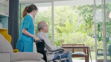 Genç Asyalı hemşire, tekerlekli sandalyede oturan yaşlı adamı destekliyor. Pencereden bahçeye bakıyor ve temiz hava, emeklilik merkezi, evdeki yaşlı adam, sağlık sigortası konseptini soluyor.