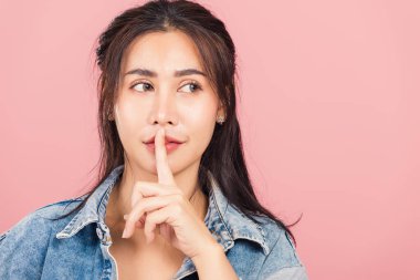 Asyalı güzel bir kadın kot pantolon giyer. Parmağını dudaklarına koyar sessiz bir jest yapar. Stüdyo resmi pembe arka planda izole edilir.