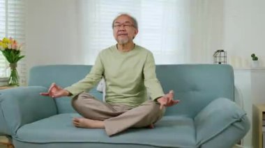 Oturma odasında kanepede oturup Mudra 'da el ele tutuşan yaşlı ve rahat bir adam, hafta sonu stresten arındıktan sonra fiziksel egzersiz yapıyor.